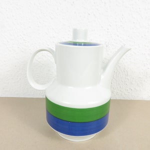 Op de afbeelding: Witte keramische koffiepot met een deksel en handvat. De pot heeft een groen en blauw horizontaal streepontwerp rond het midden. Het deksel heeft een groen en blauw streepontwerp. De pot staat op een lichtbruin oppervlak.