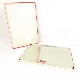 Puede incluir: Una bandeja rectangular plateada con esquinas redondeadas, dentro de una caja blanca con interior rojo. La caja está abierta, revelando la bandeja y un borde rojo. Una pequeña etiqueta roja está en la bandeja.