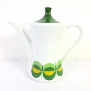 Könnte beinhalten: Weiße Keramik-Kaffeekanne mit grünem Deckel und Griff. Die Kanne hat ein dekoratives Muster aus grünen und gelben Ovalen in der Nähe des Bodens. Der Ausguss ist lang und schlank.