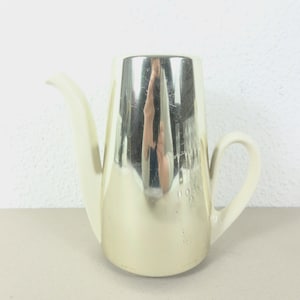 May include: A white ceramic teapot with a silver-colored metallic finish. The teapot has a curved spout and a handle.
