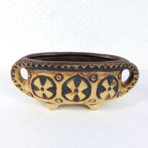 Pode incluir: Um vaso de cerâmica marrom e amarelo com três pernas e duas alças. O vaso tem um design geométrico com círculos marrons e flores estilizadas.