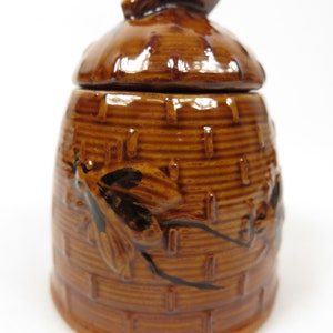 Könnte beinhalten: Ein brauner Keramik-Honigtopf in Form eines Bienenstocks mit einer Biene oben drauf. Der Topf hat eine strukturierte Oberfläche und ist mit Bienen verziert.