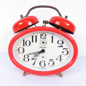 May include: A vintage red Jerger alarm clock with a white face and black numerals. The clock has a classic oval shape, two bells on top, and a metal handle. The clock face displays the time and the words "Jerger" and "Made in Germany".