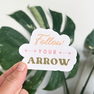 May include: A white sticker with the text "Follow Your Arrow" in gold and pink lettering. The sticker is held in front of a green leafy plant.