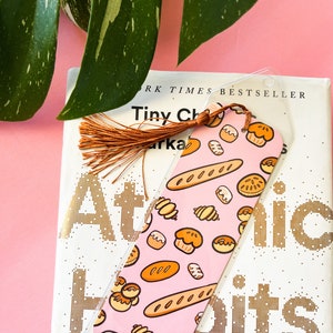 May include: A clear plastic bookmark with a pink background and a pattern of cartoon baked goods, including bread, croissants, and muffins. The bookmark has a brown tassel and rests on a book titled "Atomic Habits."