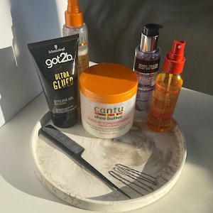 Pu&ograve; includere: Un vassoio di marmo bianco con un pettine nero, fermagli per capelli e cinque prodotti per la cura dei capelli: una bottiglia trasparente con tappo dorato, una bottiglia di gel nera e dorata, una bottiglia trasparente con tappo viola, una bottiglia trasparente con tappo rosso e un barattolo bianco con tappo arancione. Il barattolo ha il testo "Cantu shea butter leave-in conditioning repair cream" sull'etichetta.