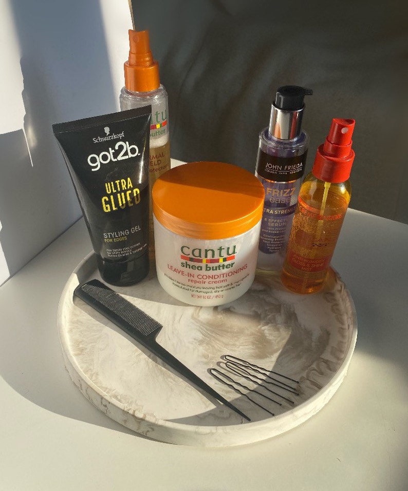 Pu&ograve; includere: Un vassoio di marmo bianco con un pettine nero, fermagli per capelli e cinque prodotti per la cura dei capelli: una bottiglia trasparente con tappo dorato, una bottiglia di gel nera e dorata, una bottiglia trasparente con tappo viola, una bottiglia trasparente con tappo rosso e un barattolo bianco con tappo arancione. Il barattolo ha il testo "Cantu shea butter leave-in conditioning repair cream" sull'etichetta.