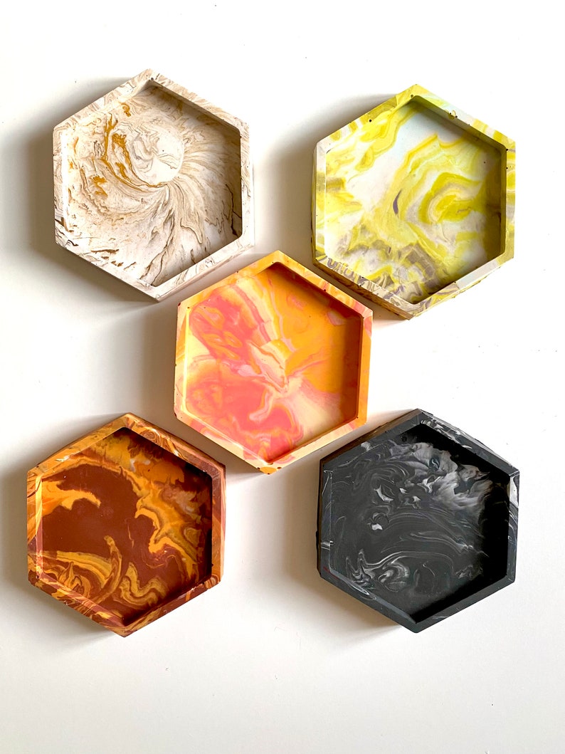 Pu&ograve; includere: Set di quattro sottobicchieri esagonali con disegni marmorei. I sottobicchieri sono in cemento e hanno un design marmoreo bianco, giallo, arancione e nero.