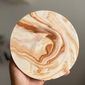 Pu&ograve; includere: Un piatto rotondo in ceramica bianca con venature marroni e beige. Il piatto ha un bordo leggermente rialzato e una finitura liscia e lucida.