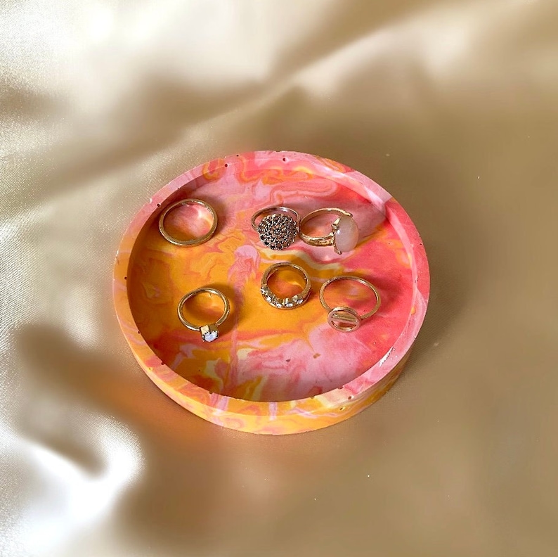 Pu&ograve; includere: Un porta anelli rotondo in cemento marmorizzato rosa e giallo con sei anelli dorati sopra.