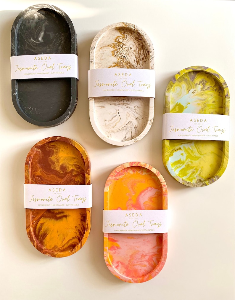 Pu&ograve; includere: Cinque vassoi ovali in jesmonite di vari colori, tra cui nero, bianco, giallo, arancione e rosa. Ogni vassoio presenta un motivo marmoreo e un'etichetta con il testo "ASEDA Jesmonite Oval Trays Handmade Homeware Sustainable".