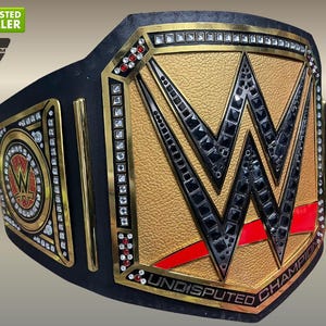 Puede incluir: Un cinturón de campeonato de lucha libre dorado y negro con el logotipo de la WWE. El cinturón presenta una placa central dorada con el logotipo de la WWE y las palabras "Undisputed Champion". El cinturón está adornado con cuero negro y detalles dorados.
