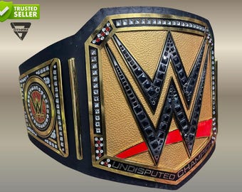 Puede incluir: Un cinturón de campeonato de cuero negro con una gran placa central dorada, un logotipo W negro prominente, detalles en rojo y cristales incrustados transparentes y rojos. La placa tiene grabado el texto UNDISPUTED CHAMPION en el borde.
