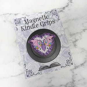 May include: A magnetic Kindle grip featuring a heart-shaped design with a purple glitter border and a central illustration of a bat with pink wings against a rainbow background. The product is mounted on a card labelled Magnetic Kindle Grips.