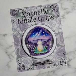 May include: A circular magnetic Kindle grip featuring a mushroom illustration with a purple and blue galaxy cap, a white stem, and a yellow crescent moon. The grip is mounted on a rectangular display card with the text Magnetic Kindle Grips.