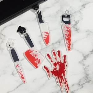May include: A collection of acrylic charms featuring a horror theme. The charms include a knife, cleaver, saw, and a hand, all with red blood splatter details. A black Kindle device is in the background.