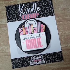 May include: A black circular Kindle Grip phone accessory featuring a white rectangular graphic of colourful books with hearts. The text reads Kindle GRIP, Bookish GIRLIE, and Literary FILTH. The item is mounted on a black and white book-patterned card.