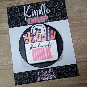 May include: A circular black phone grip featuring a white rectangular graphic with a row of colourful books in shades of pink, orange, and yellow. The text reads Bookish GIRLIE in black and pink script and block letters, with small pink hearts.