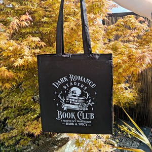 May include: Black tote bag with the text "Dark Romance Readers Book Club" in white. The design includes a skull, books, and a rose. The bag has long handles and is displayed against a backdrop of yellow and green foliage.