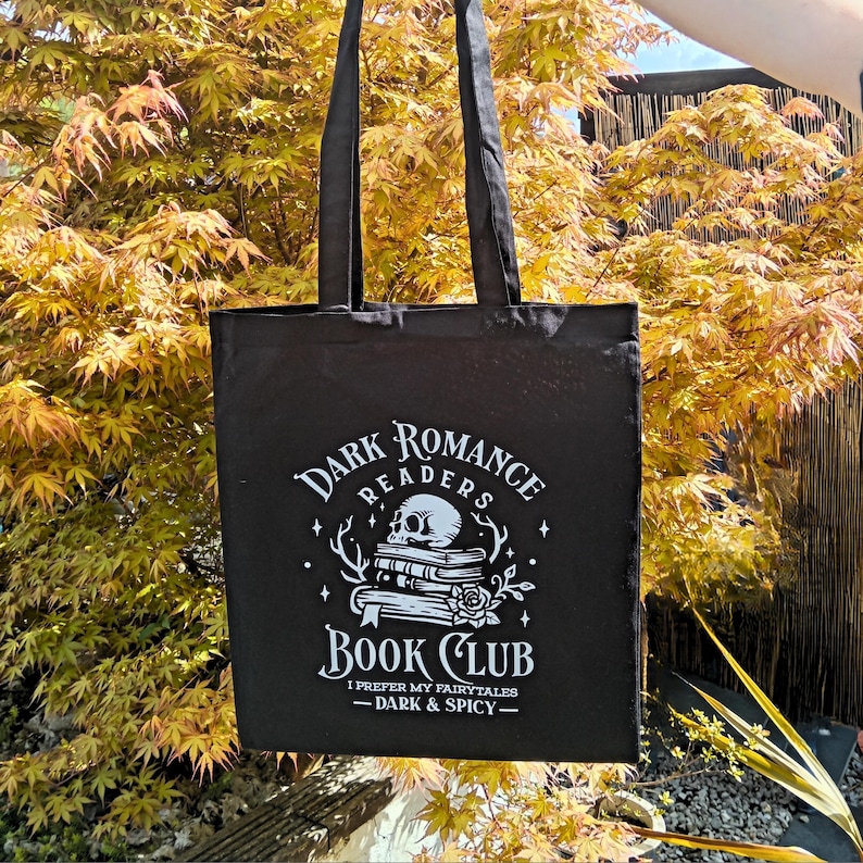 May include: Black tote bag with the text "Dark Romance Readers Book Club" in white. The design includes a skull, books, and a rose. The bag has long handles and is displayed against a backdrop of yellow and green foliage.