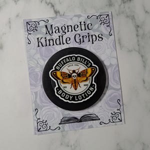 May include: A black magnetic Kindle grip with a circular design featuring a moth with a skull on a white background. The text "BUFFALO BILL'S BODY LOTION" and "Magnetic Kindle Grips" are also visible. The background is light grey with a floral pattern.