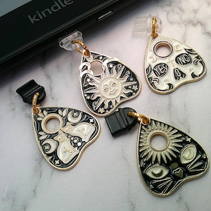May include: Four black, white, and gold-coloured Ouija planchette charms with various designs. One features a smiling sun, another has a skull with "YES" and "NO" text, and the others have cat and moon designs. Each charm has a black clasp.