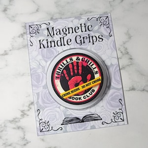 May include: A circular magnetic Kindle grip on a display card labelled Magnetic Kindle Grips. The grip features a red handprint, the text Thrills & Chills Book Club, and a yellow crime scene tape graphic with the text CRIME SCENE - DO NOT CROSS.