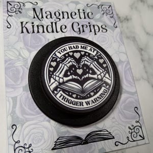 May include: A black magnetic Kindle grip with the text "YOU HAD ME AT TRIGGER WARNING". The design features skeleton hands forming a heart over an open book, with hearts and stars. The grip is on a card with the text "Magnetic Kindle Grips".