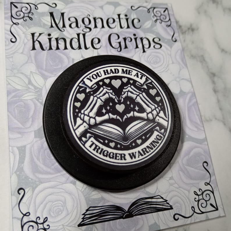 May include: A black magnetic Kindle grip with the text "YOU HAD ME AT TRIGGER WARNING". The design features skeleton hands forming a heart over an open book, with hearts and stars. The grip is on a card with the text "Magnetic Kindle Grips".