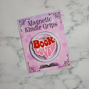 May include: A circular magnetic Kindle grip featuring a sticker with the text BOOK LOVER in red and pink retro-style lettering. The grip is mounted on a rectangular pink display card with heart patterns and the text Magnetic Kindle Grips.