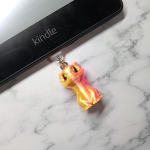 May include: A vibrant, 3D-printed dragon charm in yellow, orange, and pink. The dragon is connected to a black Kindle device with a gold chain. The word "kindle" is printed on the device.