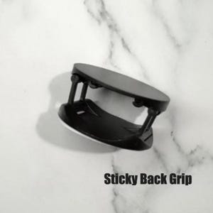 May include: A black sticky back grip for a phone or tablet. The grip has a circular base and a rectangular top, connected by two black supports. The text "Sticky Back Grip" is printed in bold, black letters.