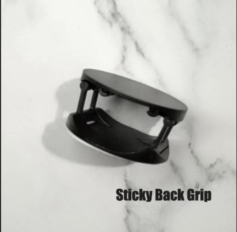 May include: A black sticky back grip for a phone or tablet. The grip has a circular base and a rectangular top, connected by two black supports. The text "Sticky Back Grip" is printed in bold, black letters.