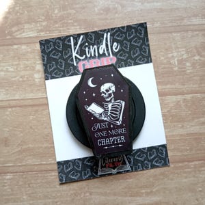 May include: A black coffin-shaped phone grip featuring a white skeleton reading a book under a crescent moon and stars. The text reads "JUST ONE MORE CHAPTER". It is mounted on a white display card with the text "Kindle GRIP" and "Literary FILTH".