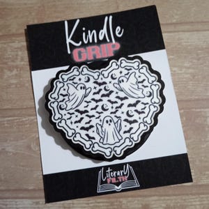 May include: A heart-shaped Kindle Grip accessory featuring a white background with a black pattern of ghosts, bats, moons, and stars, bordered by a ring of white bones. The packaging displays the text Kindle GRIP and Literary FILTH.