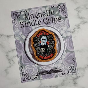 May include: A magnetic Kindle grip with the text "JUST THE TIP I PROMISE" and a horror movie character image. The grip is orange and glittery, set against a floral background. The card reads "Magnetic Kindle Grips".