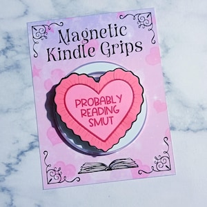 May include: A pink and white magnetic Kindle grip with a heart-shaped design. The heart has a ruffled edge and the text "PROBABLY READING SMUT". The card reads "Magnetic Kindle Grips" with decorative flourishes and a book illustration.