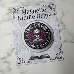 May include: A circular magnetic Kindle grip featuring a graphic of a skull resting on an open book surrounded by purple roses. The text reads Dark Romance Book Club. It is mounted on a rectangular display card with a rose pattern and the text Magnetic Kindle Grips.