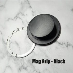 May include: A black Mag Grip with a circular design, featuring a raised center and a white circular sticker. The text "Mag Grip - Black" is displayed at the bottom of the image. The background is a white marble pattern.