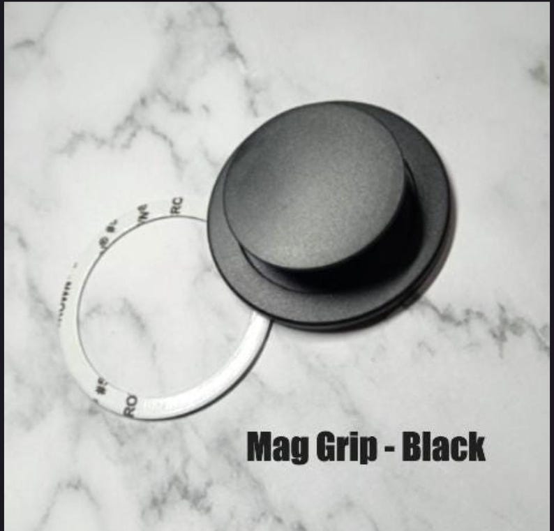 May include: A black Mag Grip with a circular design, featuring a raised center and a white circular sticker. The text "Mag Grip - Black" is displayed at the bottom of the image. The background is a white marble pattern.