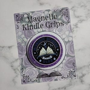 May include: A circular purple glitter magnetic grip on a backing card labelled Magnetic Kindle Grips. The grip features a black centre with a white open book illustration, a crescent moon, stars, and the text Midnight Readers Club One More Chapter.