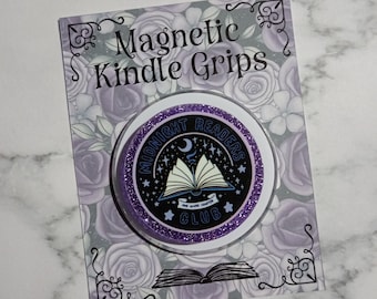 Può includere: Un'impugnatura magnetica circolare con glitter viola su un cartoncino con l'etichetta Magnetic Kindle Grips. L'impugnatura presenta un centro nero con un'illustrazione di un libro aperto bianco, una luna crescente, stelle e il testo Midnight Readers Club One More Chapter.