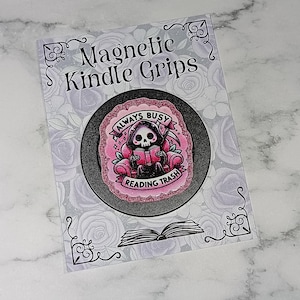 May include: A circular black magnetic Kindle grip featuring a pink glittery illustration of a hooded skeleton reading a book in a chair. The text reads Always Busy Reading Trash. It is mounted on a rectangular card labelled Magnetic Kindle Grips.