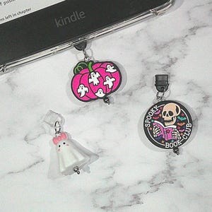 May include: A selection of Halloween-themed charms, including a pink pumpkin with white ghosts, a white ghost with a pink bow, and a circular charm with a skull reading a book. The charms are attached to a Kindle.
