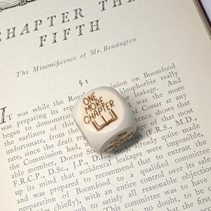 May include: A white die with the text "ONE MORE CHAPTER" and a book illustration, placed on an open book. The book's page shows the title "CHAPTER THE FIFTH".
