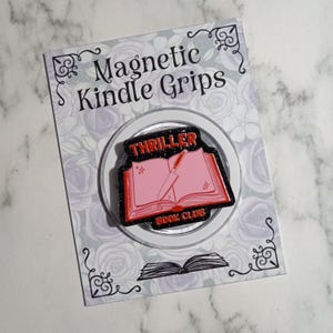May include: A circular magnetic Kindle grip featuring an enamel pin design of an open book with a knife resting on the pages. The text reads THRILLER BOOK CLUB in red. The background is black with glitter, mounted on a card labelled Magnetic Kindle Grips.
