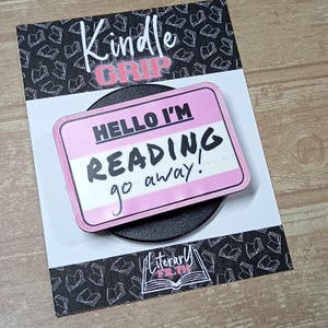 May include: A rectangular pink and white name tag accessory with black text that reads HELLO I'M READING go away. The accessory is mounted on a black circular base and attached to a white display card featuring a black book pattern and text.