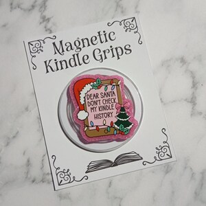 May include: A pink glitter magnetic Kindle grip with a festive design. The design features a Santa hat, Christmas tree, and the text "DEAR SANTA DON'T CHECK MY KINDLE HISTORY". The grip is on a white card with the text "Magnetic Kindle Grips".