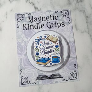 May include: A magnetic Kindle grip with a design featuring an open book, glasses, a coffee cup, and the text "Just one more chapter." The grip is on a decorative card with the words "Magnetic Kindle Grips".