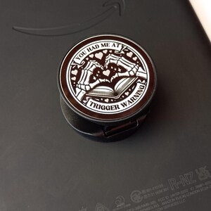 May include: A black and brown phone grip with the text "YOU HAD ME AT TRIGGER WARNING" in a circular design. The design includes skeleton hands, hearts, and an open book. The grip is attached to a black electronic device.